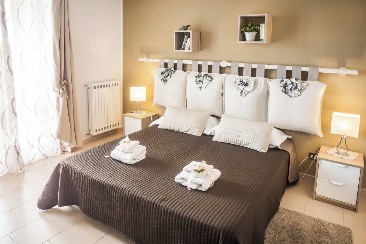 A neatly made bed with a brown bedspread, two white pillows, and two folded towels on the bed. There are two bedside tables with lamps, a wooden headboard, and a shelf above the bed with books and a potted plant.
