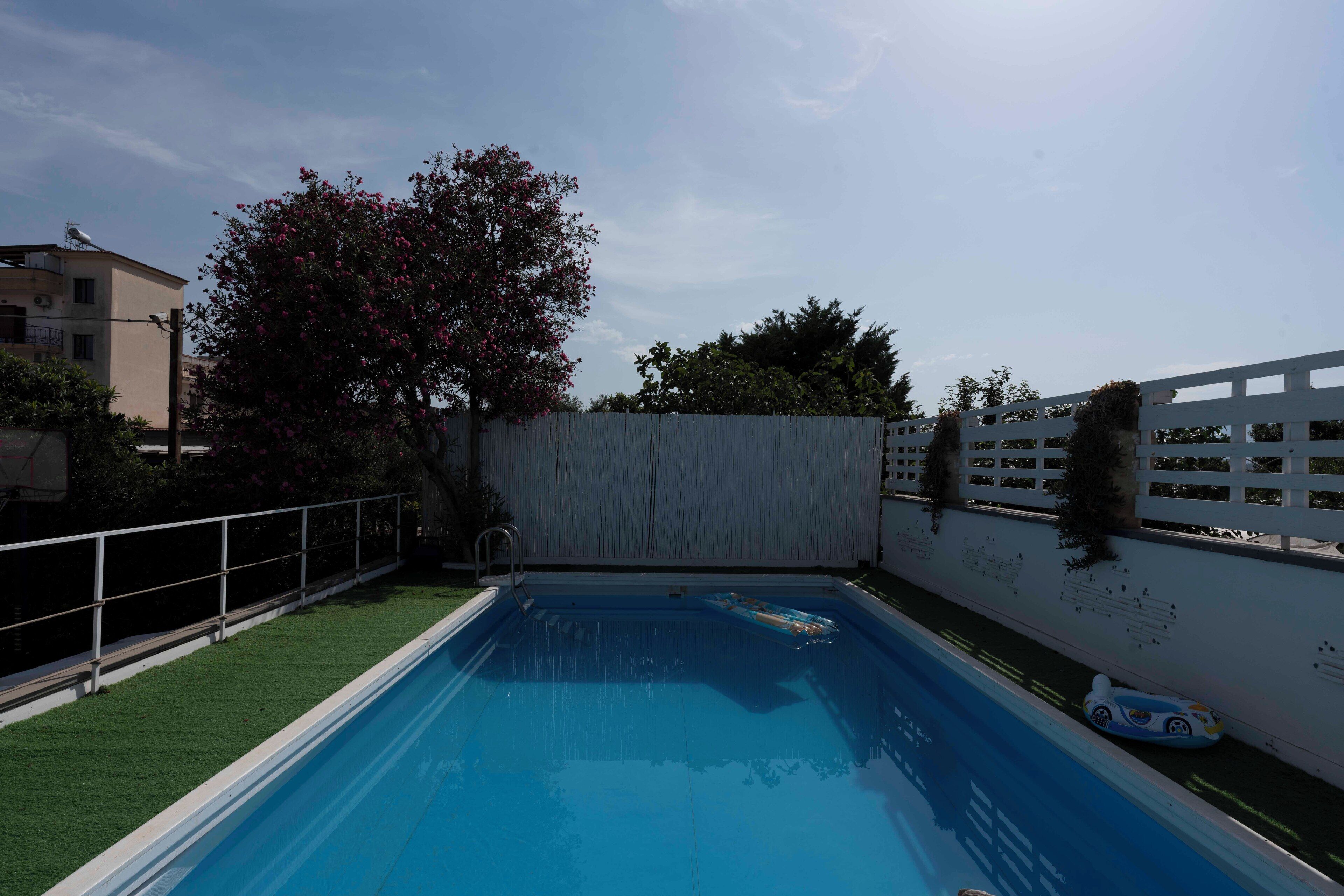 Una piscina rectangular con superficie de agua azul cristalina, rodeada por una cerca blanca y un árbol con flores rosadas.