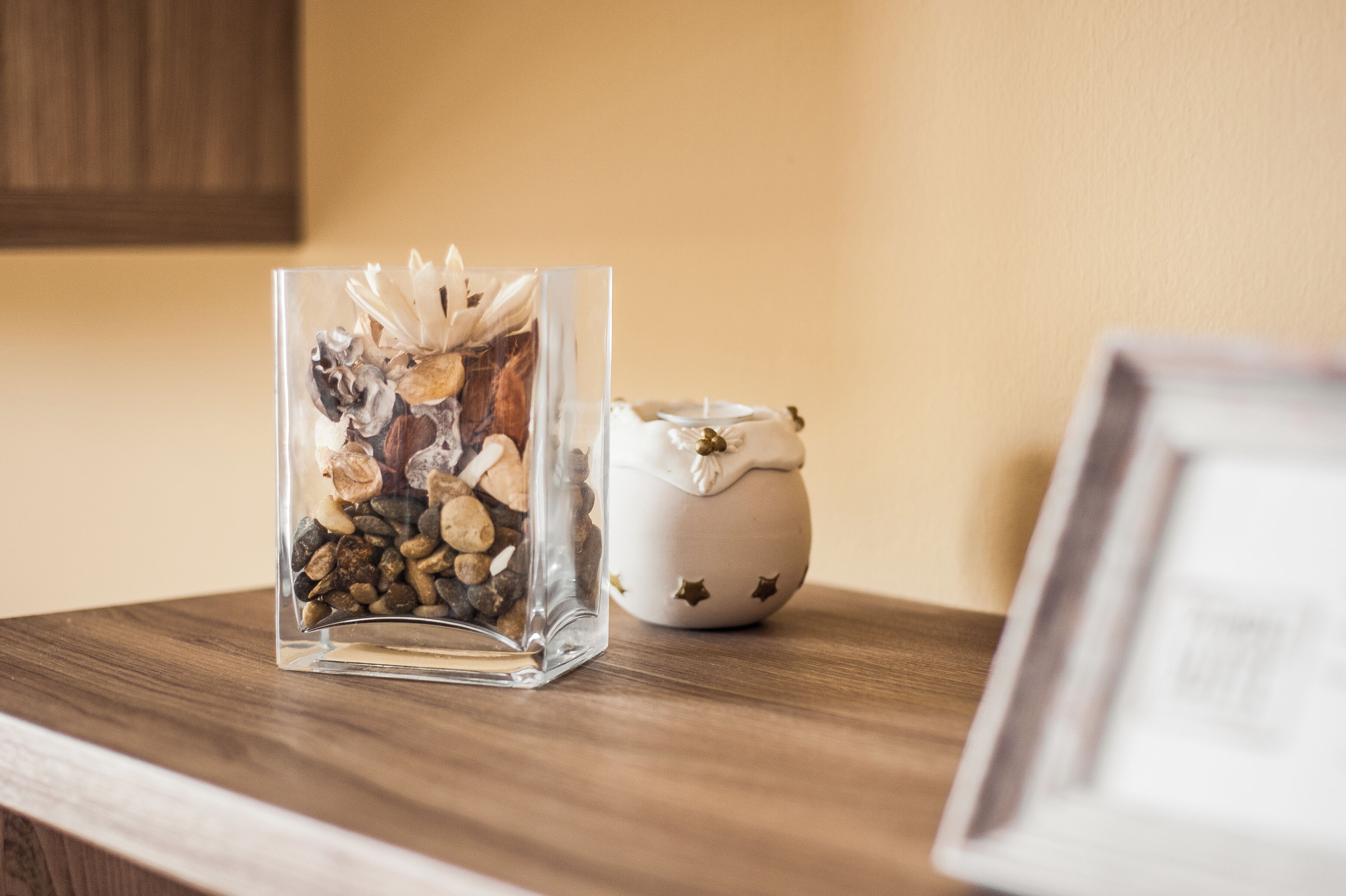 Un vase en verre transparent contenant des fleurs séchées et des petits galets, posé sur une surface en bois, à côté d’une tasse en céramique ornée de motifs en forme d’étoiles.