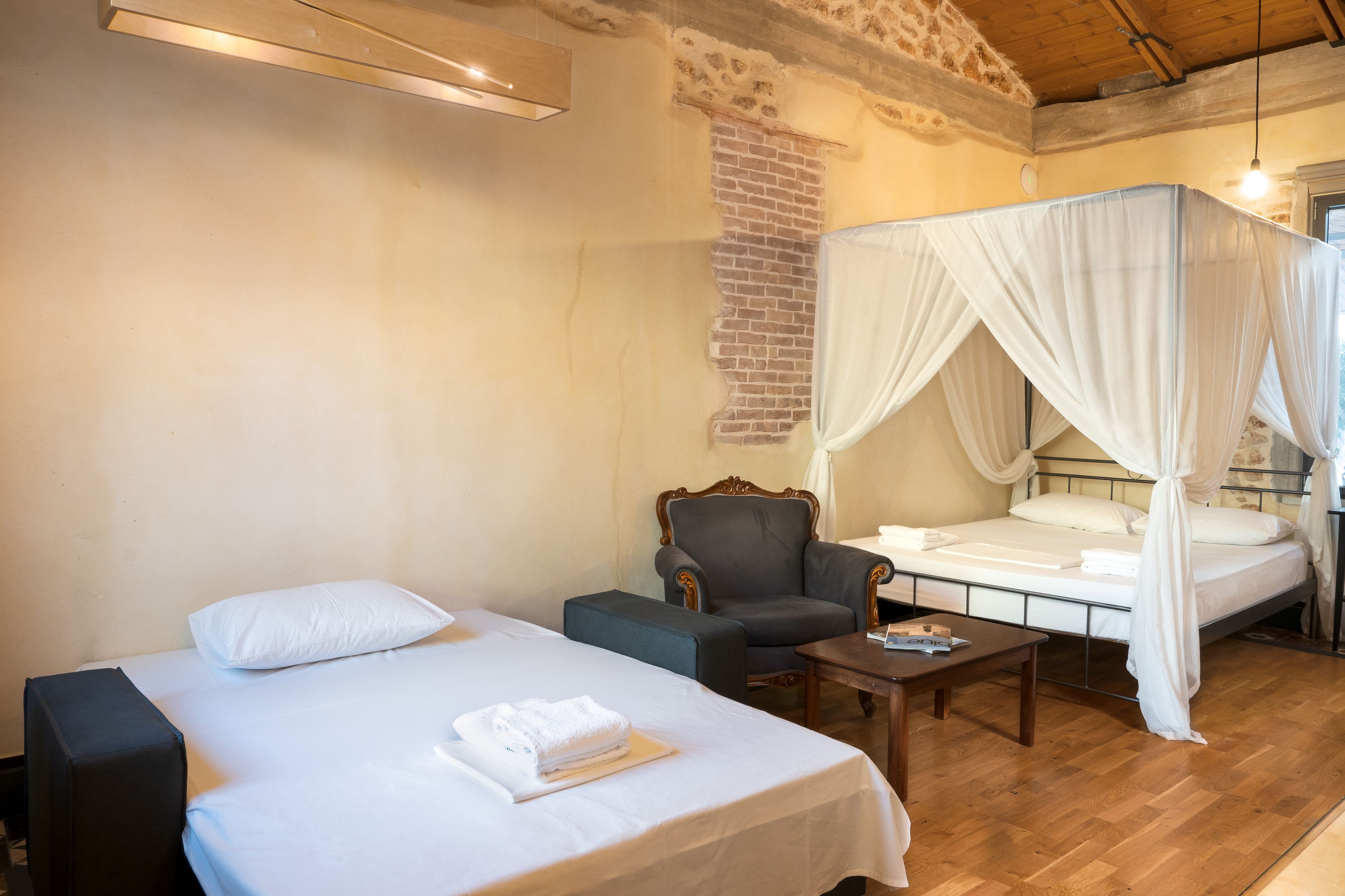 A bedroom with two beds, a chair, a table, and a brick wall.