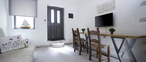 A small, clean room with a sofa, a dining table with chairs, a TV mounted on the wall, and a window with a sailboat decoration.