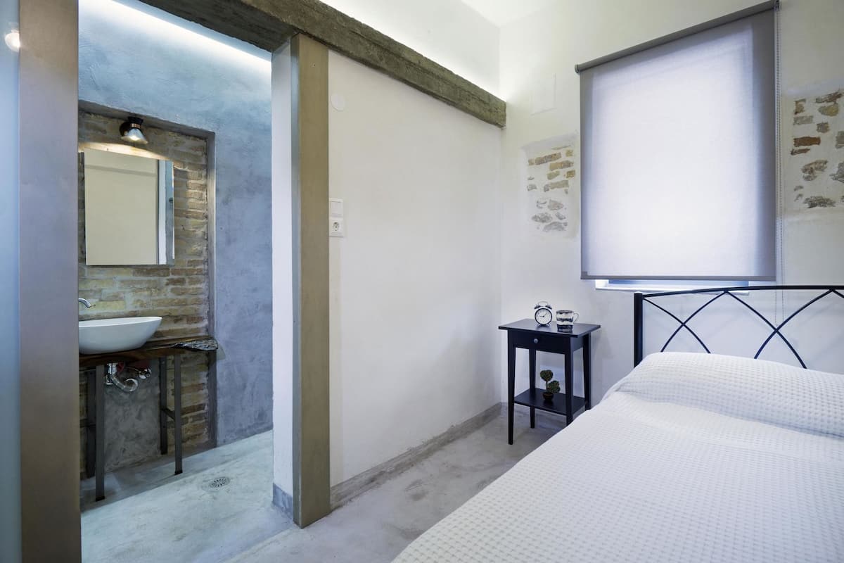 Ein Schlafzimmer mit Bett, Nachttisch mit Uhr und Tasse, ein Badezimmer mit Waschbecken und Spiegel sowie ein Fenster mit Blick zum Himmel.