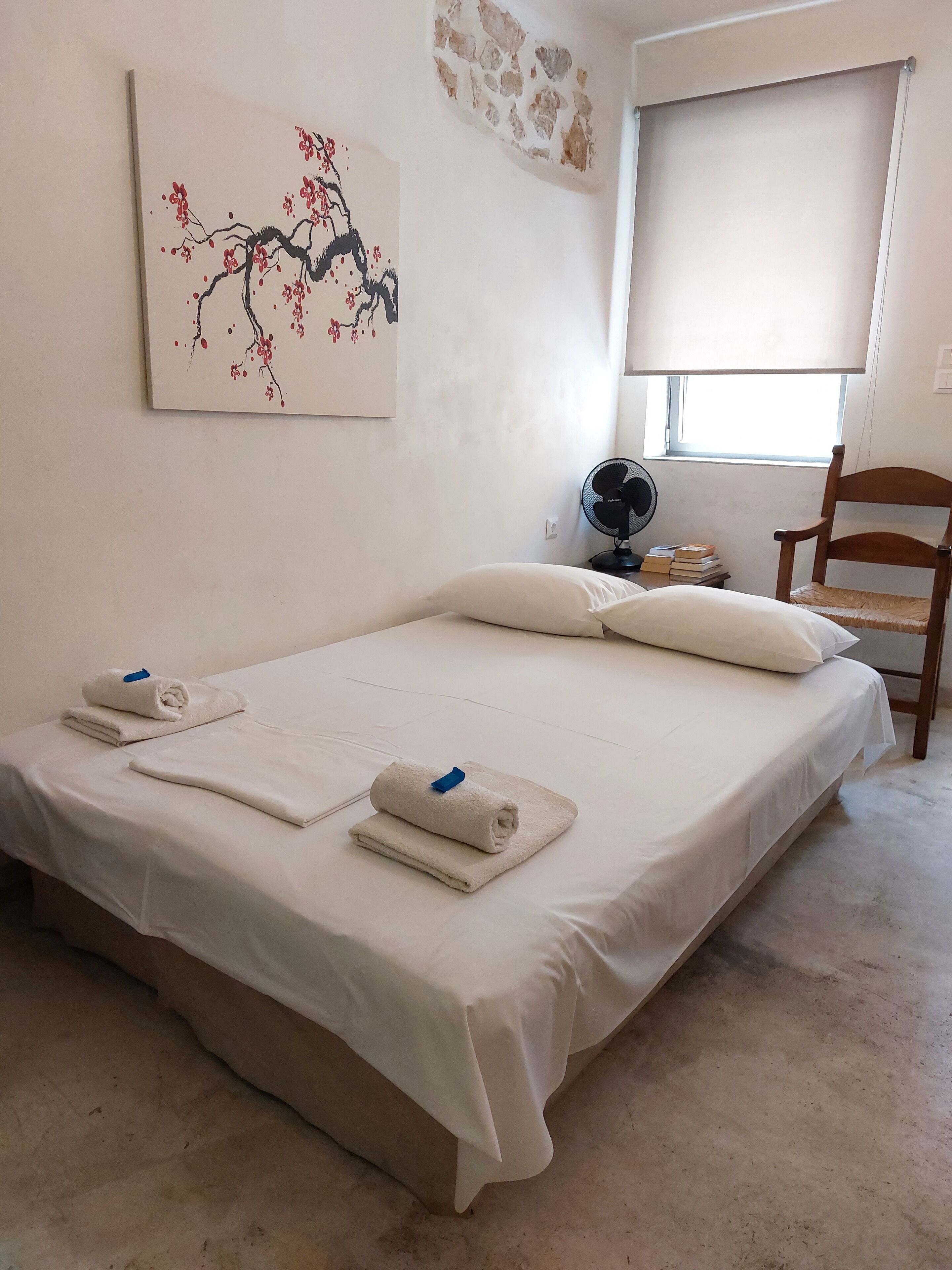 A neatly made bed with white linens and towels, a wooden chair, a fan, and a window with a view.