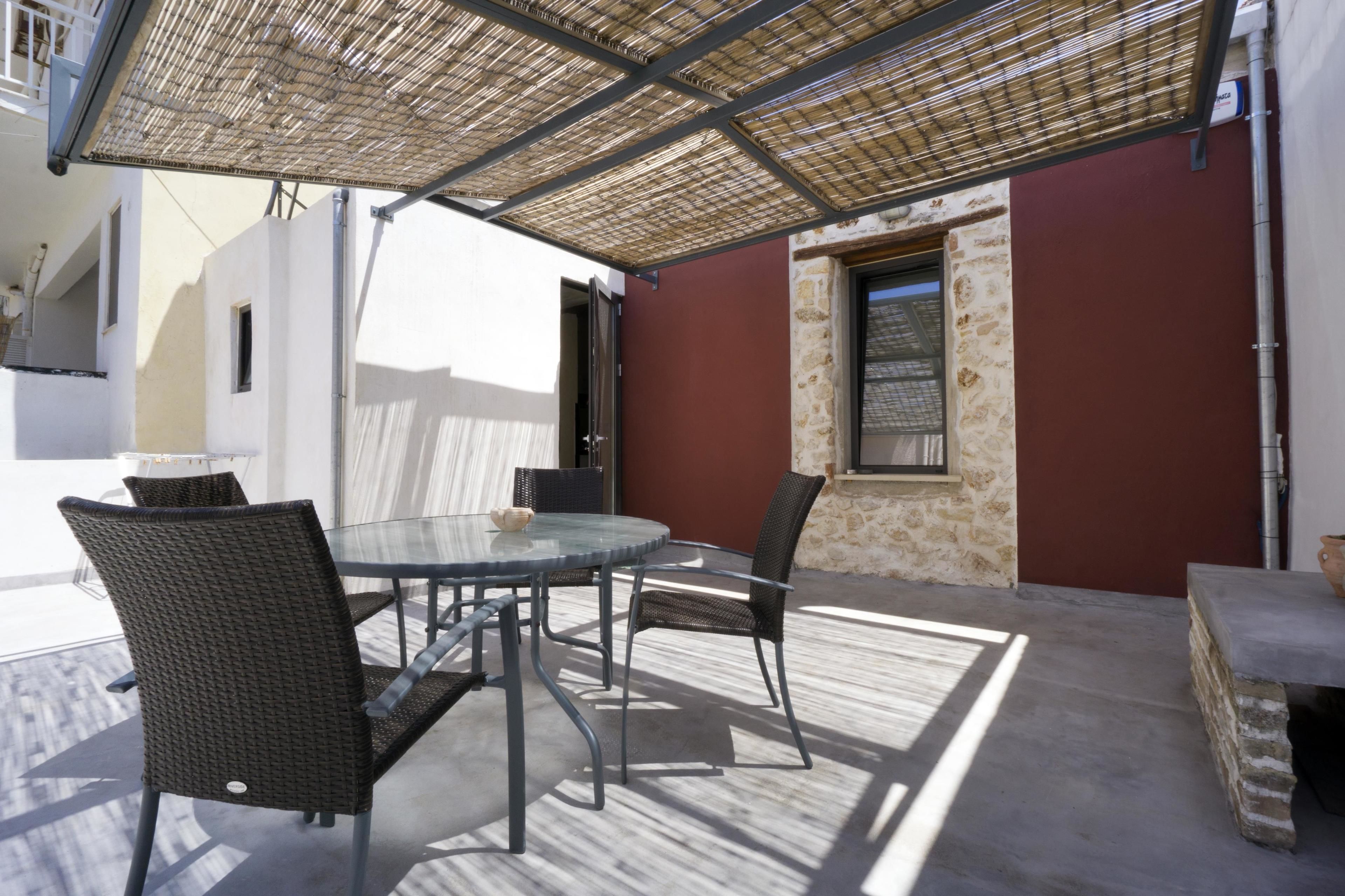 Un patio al aire libre con una mesa de vidrio y sillas de mimbre, un muro de piedra y una pérgola de metal que proporciona sombra.
