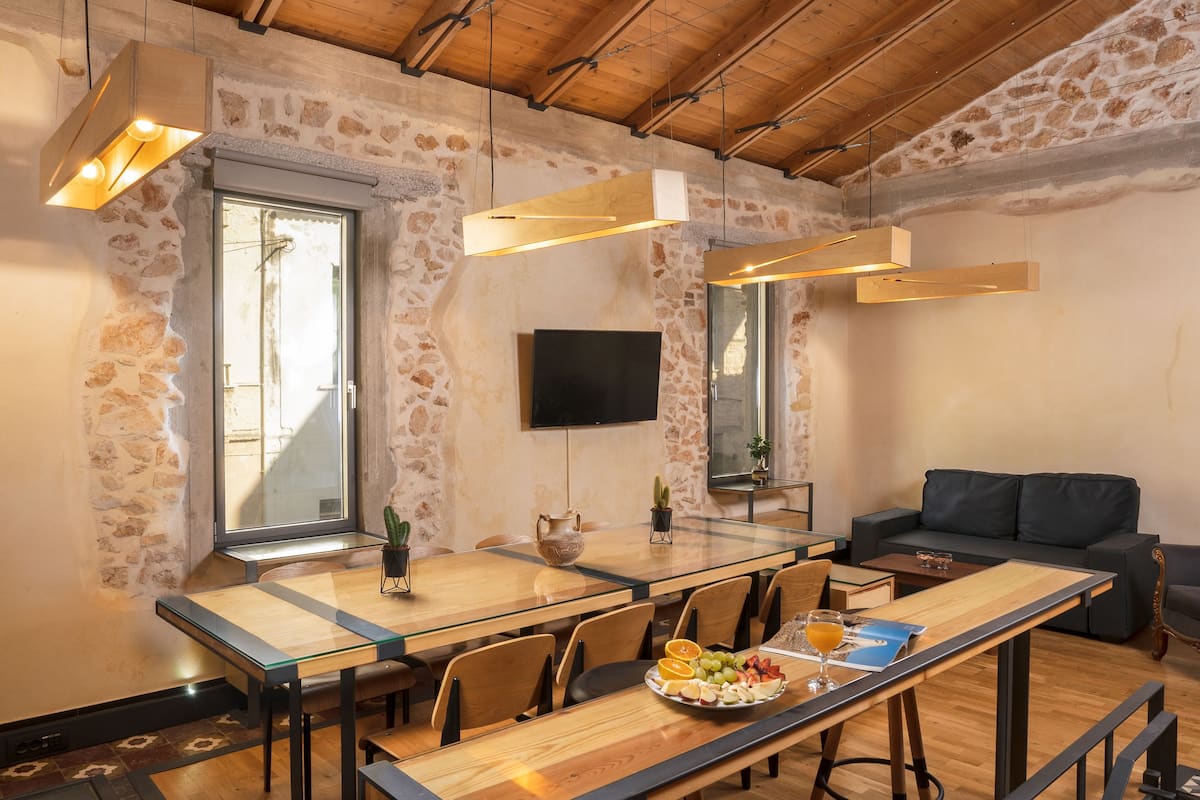 Ein moderner Essbereich mit einem langen Tisch, Stühlen und einer Glastrennwand. An der Wand ist ein Flachbildfernseher angebracht, und im Hintergrund steht eine Couch.