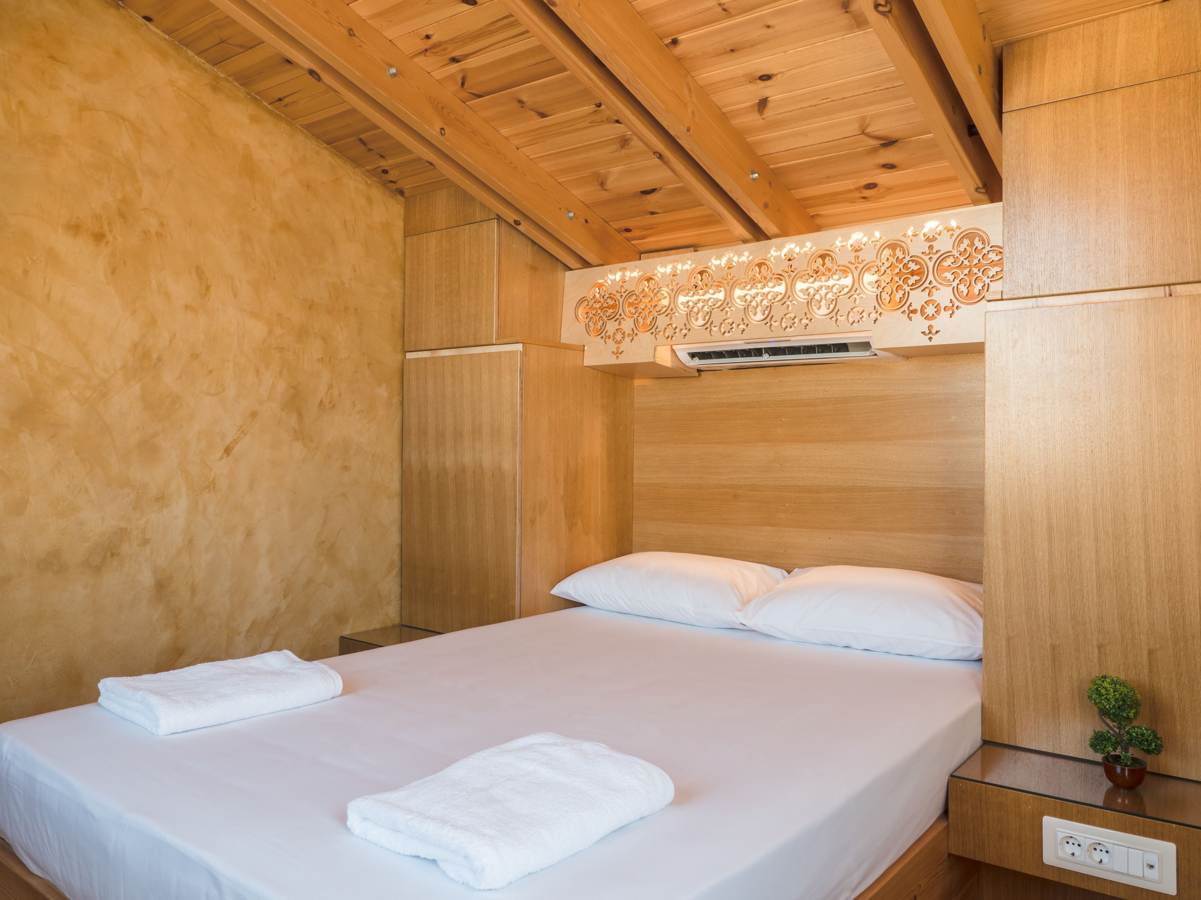 A hotel room with a wooden ceiling, a bed with white linens, a small bedside table with a plant, and a wall-mounted air conditioning unit.