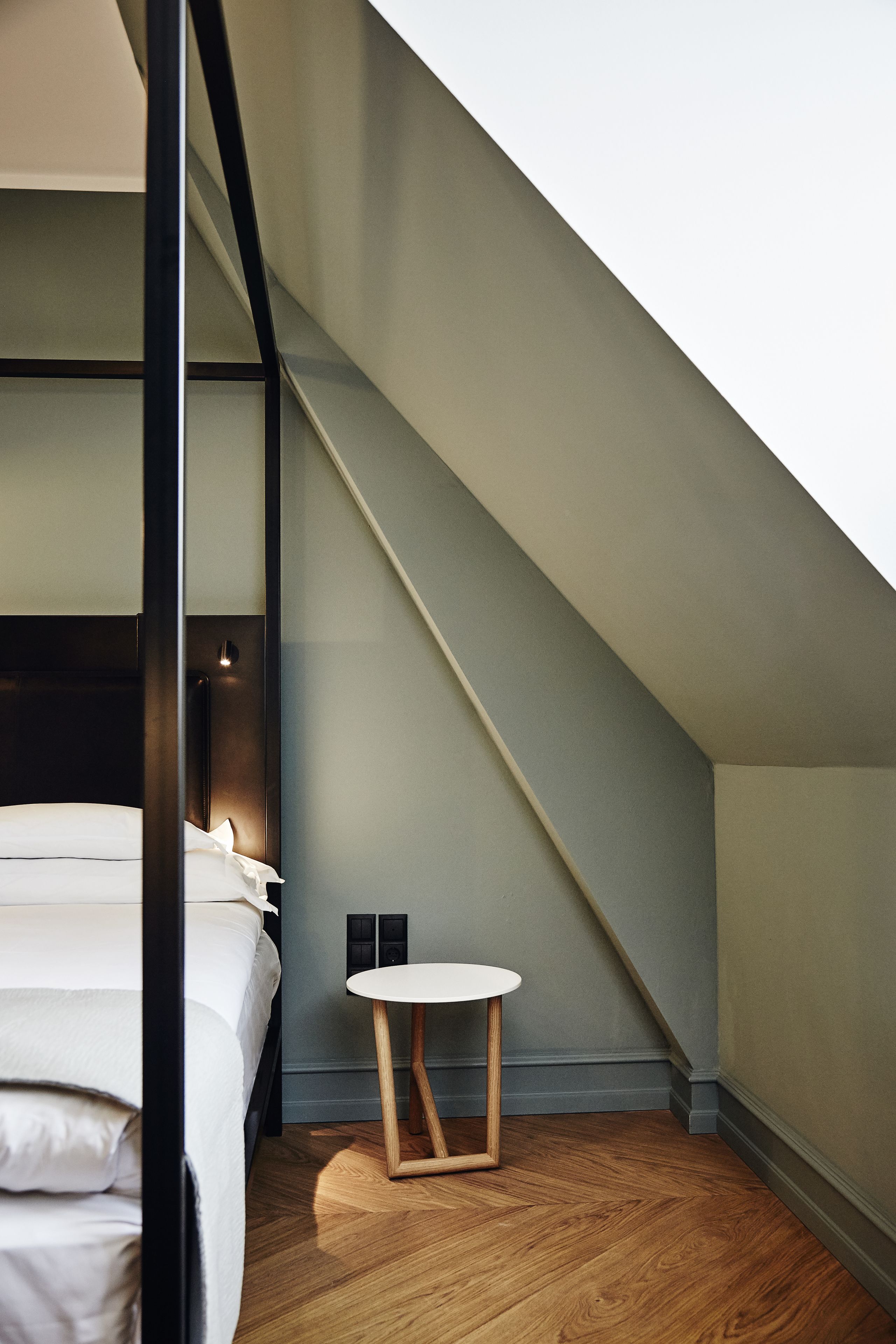 Un dormitorio moderno con techo inclinado, una cama con ropa de cama blanca, una mesita redonda de madera y una pequeña lámpara fijada a la pared.