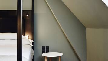 Un dormitorio moderno con techo inclinado, una cama con ropa de cama blanca, una mesita redonda de madera y una pequeña lámpara fijada a la pared.