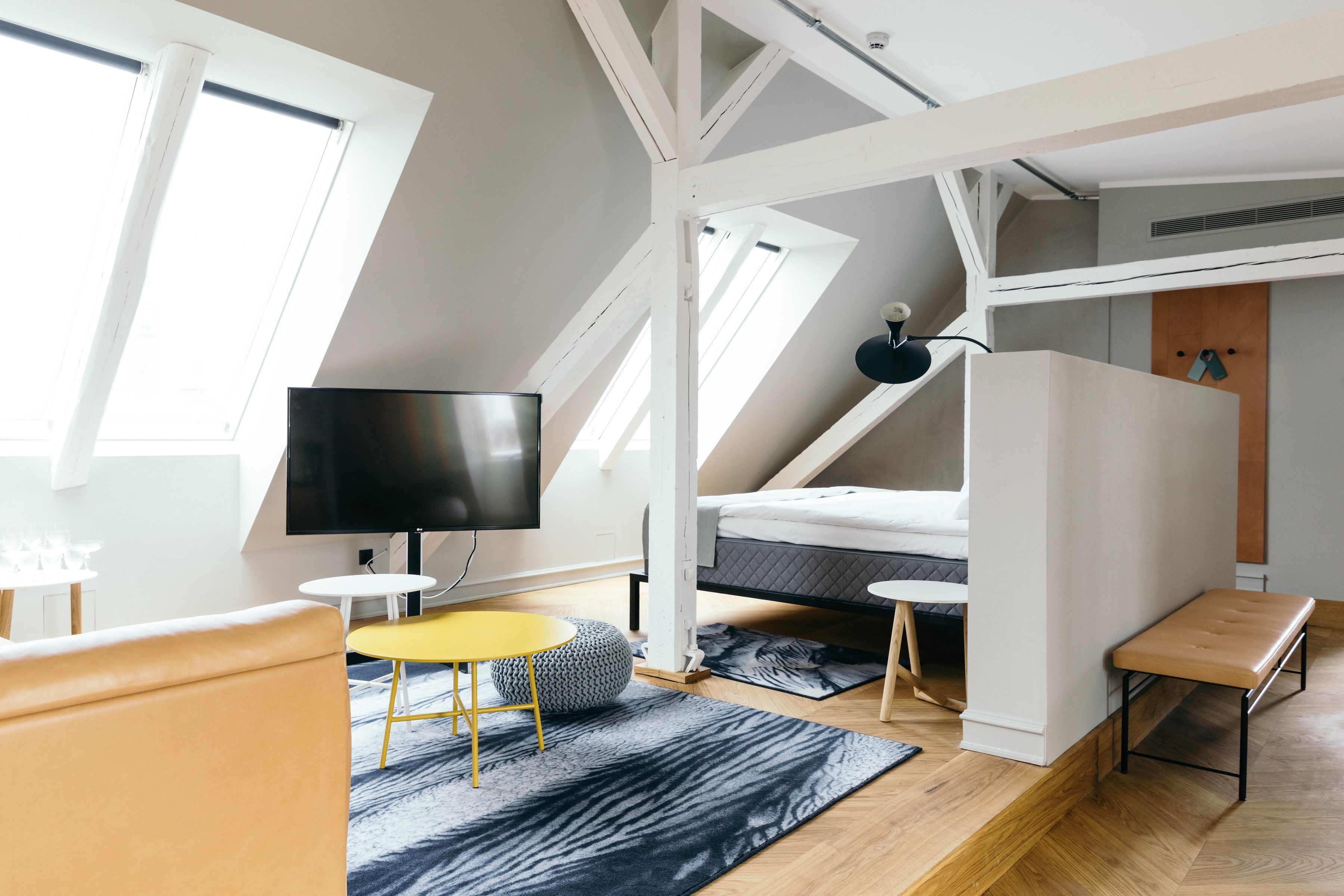 A modern loft bedroom with a sloped ceiling, a bed, a TV, and a small seating area with a yellow table and ottoman.