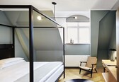 A modern hotel room with a four-poster bed, a desk with a chair, and a window with a view.