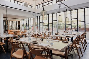 Un comedor amplio con techos altos, ventanas grandes y modernas sillas de madera. Las mesas están dispuestas con platos, vasos y cubiertos.