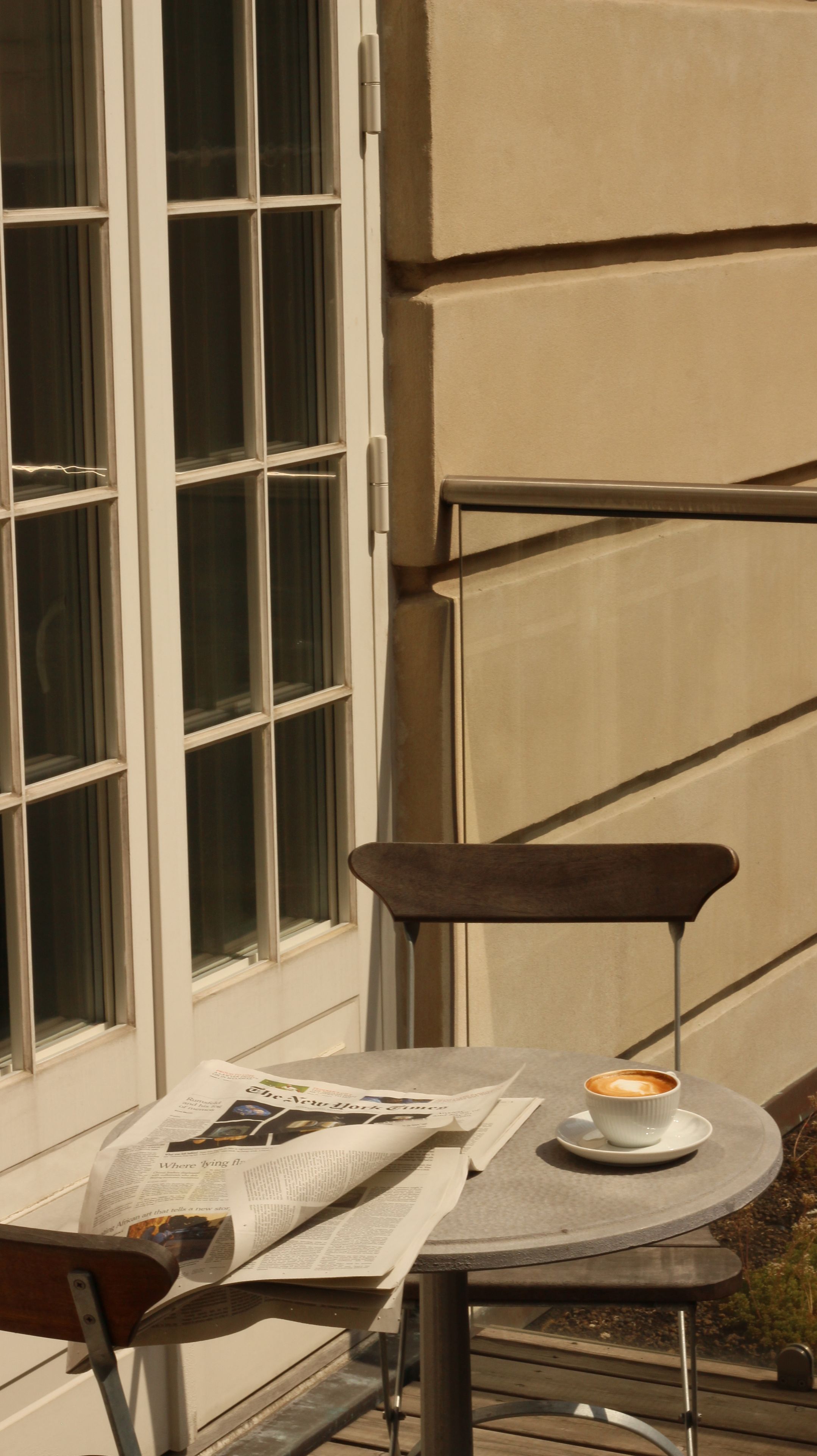 A table with a newspaper and a cup of coffee, a chair, and a window with blinds.