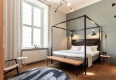 A modern bedroom with a four-poster bed, a bench, a small table, and a large window with curtains.