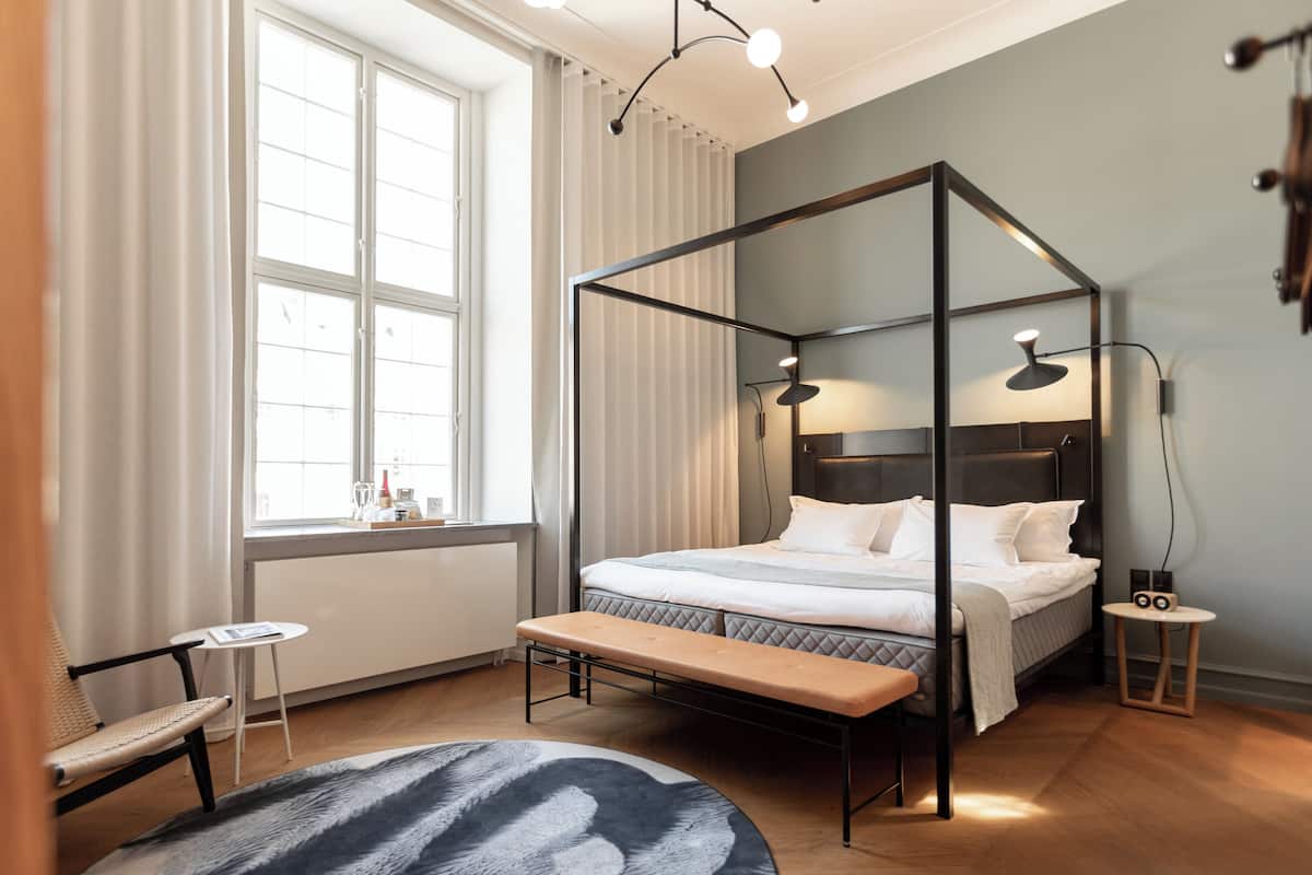 Une chambre à coucher moderne avec un lit à baldaquin, un banc, une petite table et une grande fenêtre avec des rideaux.