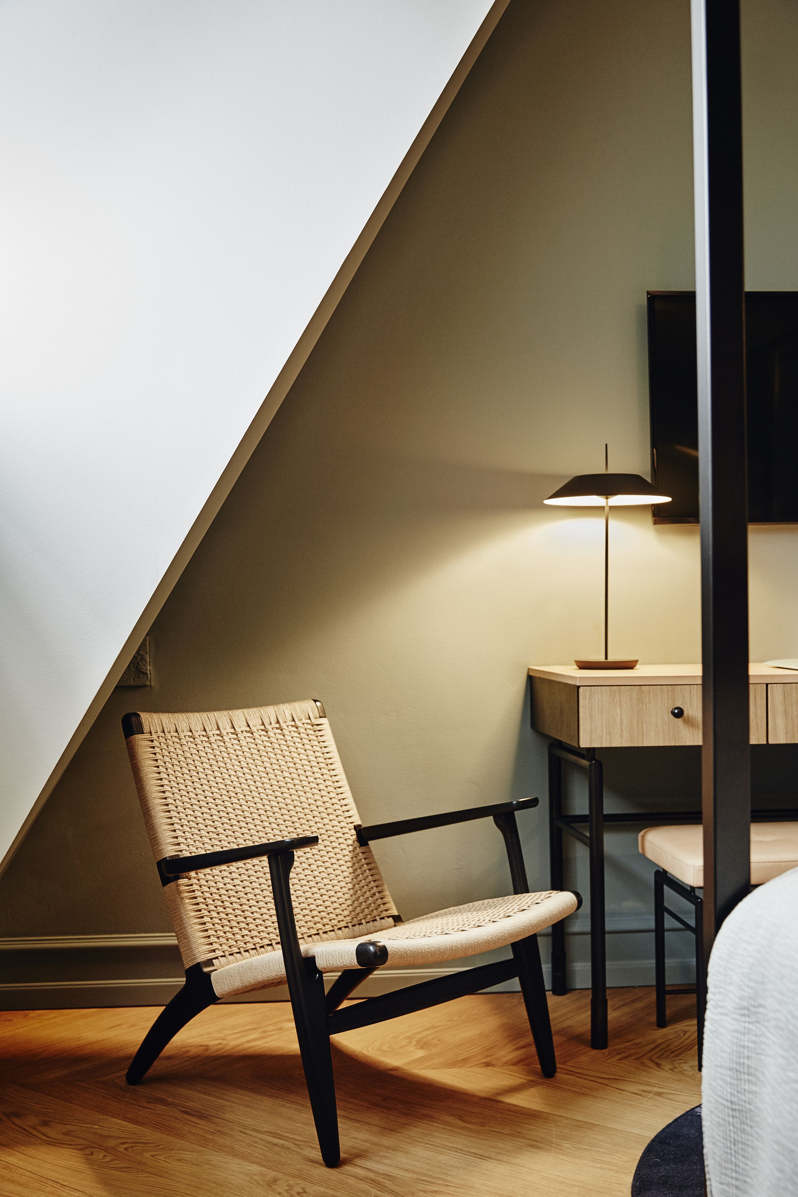 A modern room with a sloped ceiling, a woven chair, a desk with a lamp, and a television.