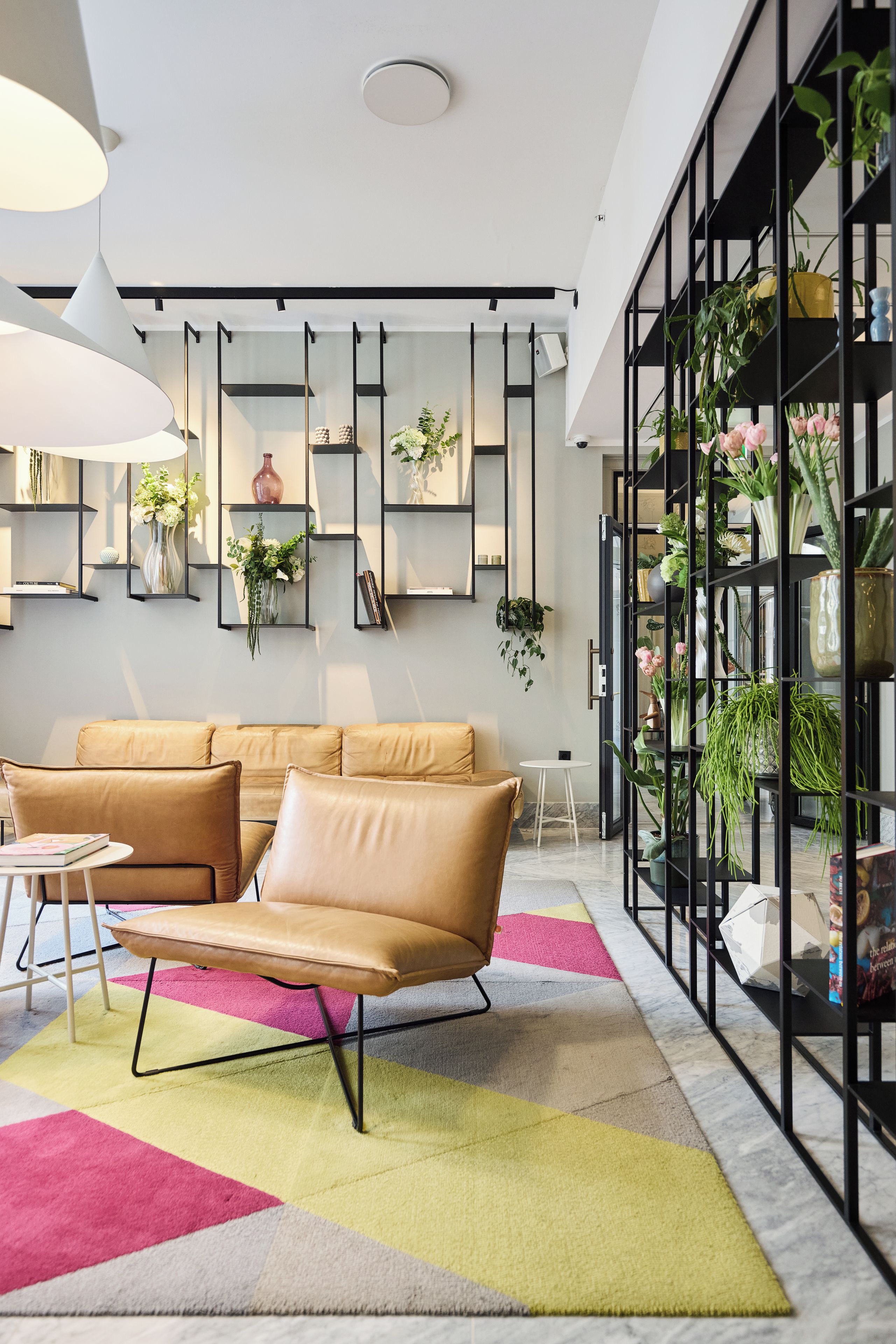 A modern living room with a beige sofa, a round coffee table, and a colorful rug. There are shelves with plants and decorative items, and a large window with a view of the outdoors.