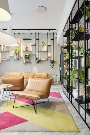 A modern living room with a beige sofa, a round coffee table, and a colorful rug. There are shelves with plants and decorative items, and a large window with a view of the outdoors.