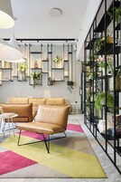 Una sala moderna con un sofá beige, una mesa redonda y una alfombra colorida. Hay estanterías con plantas y objetos decorativos, y un ventanal con vistas al exterior.