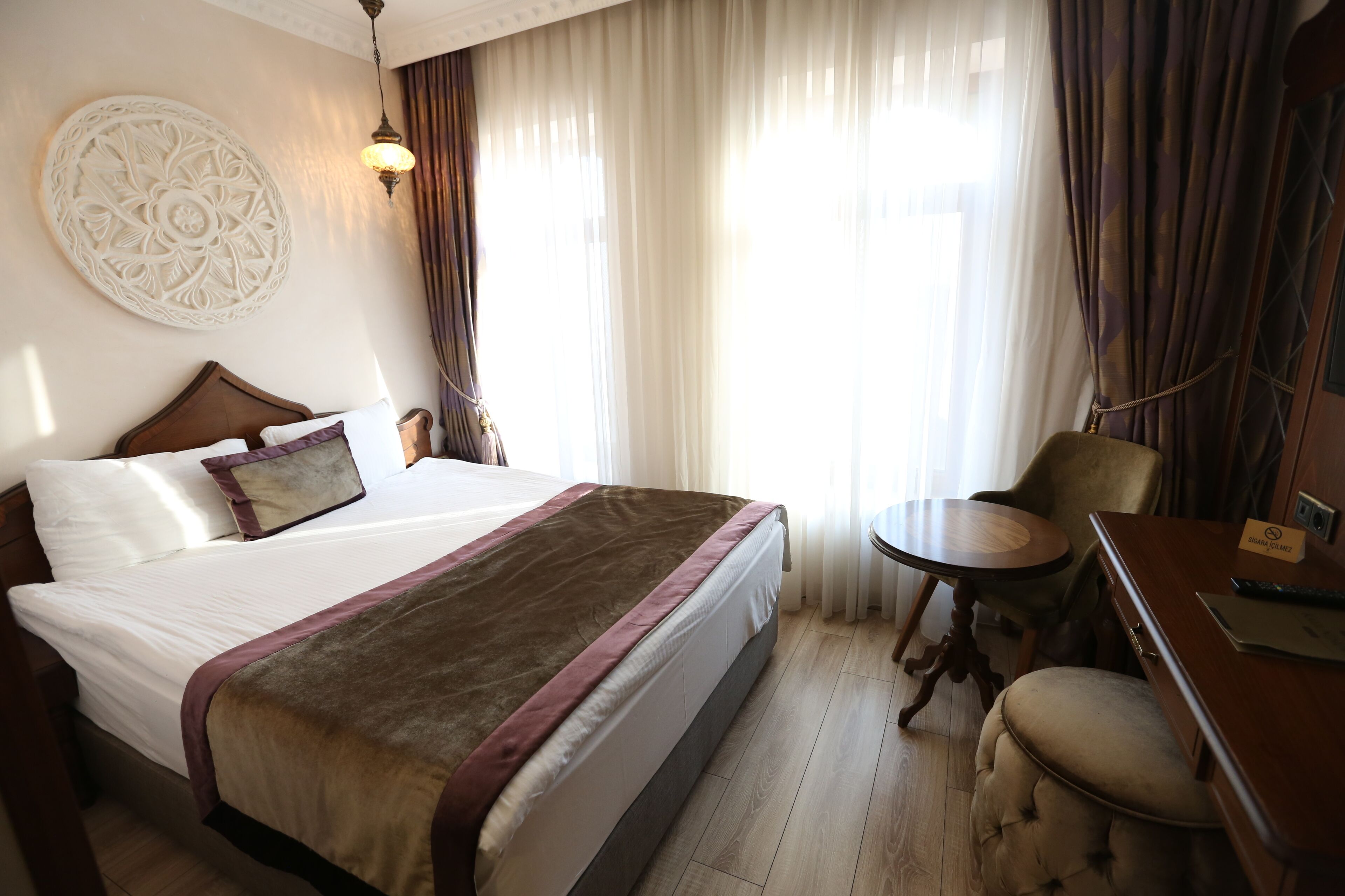 Quarto de hotel com uma cama grande, uma mesa redonda pequena, uma cadeira e uma escrivaninha com um computador e uma placa.