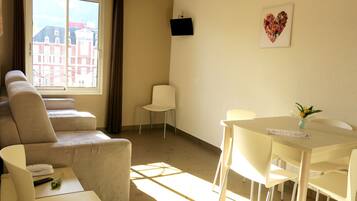 A room with a sofa, a dining table with chairs, a small white table, a TV mounted on the wall, and a window with a view of buildings.