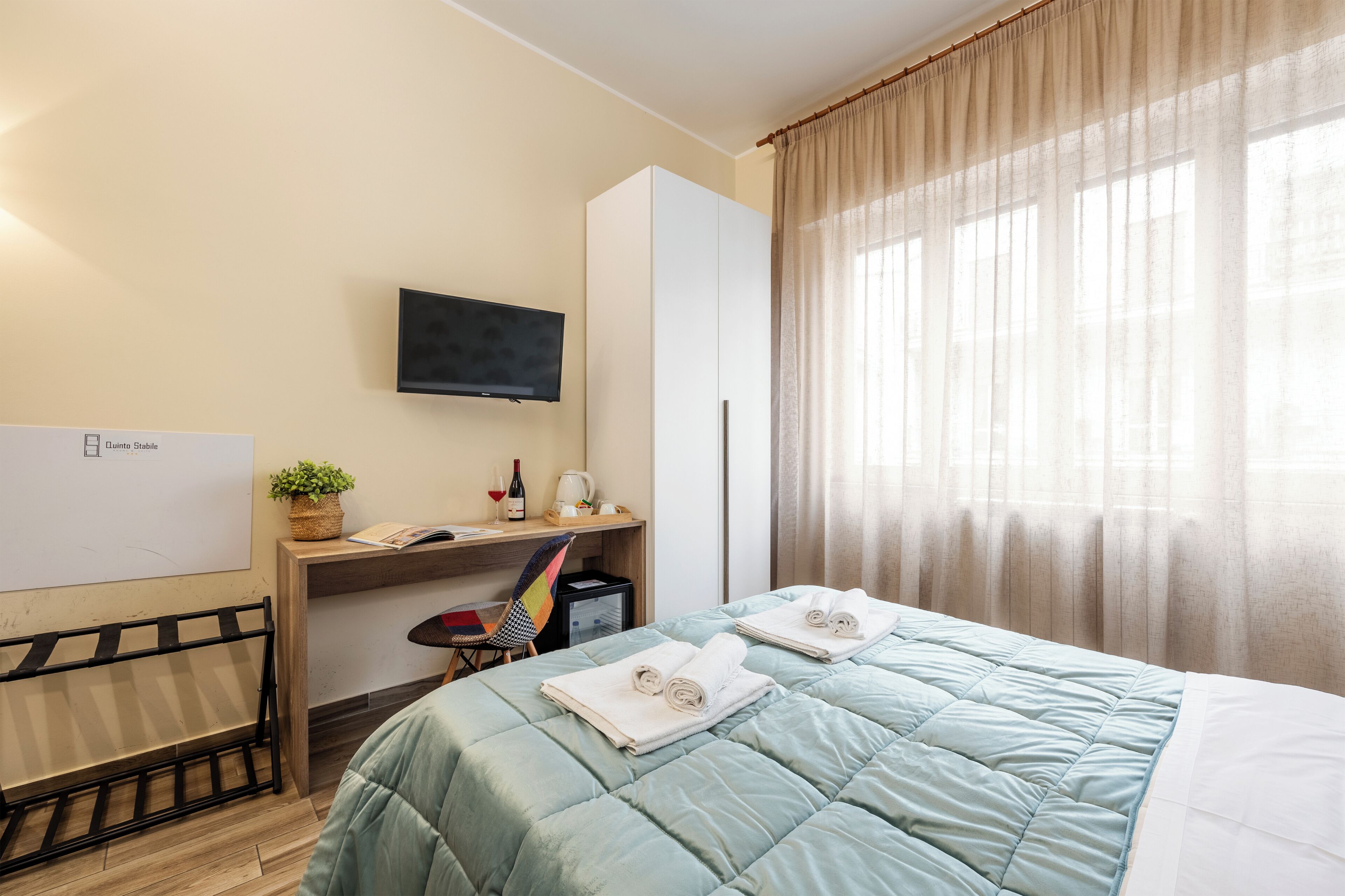 A hotel room with a bed, a desk, a chair, a TV, and a window with curtains.