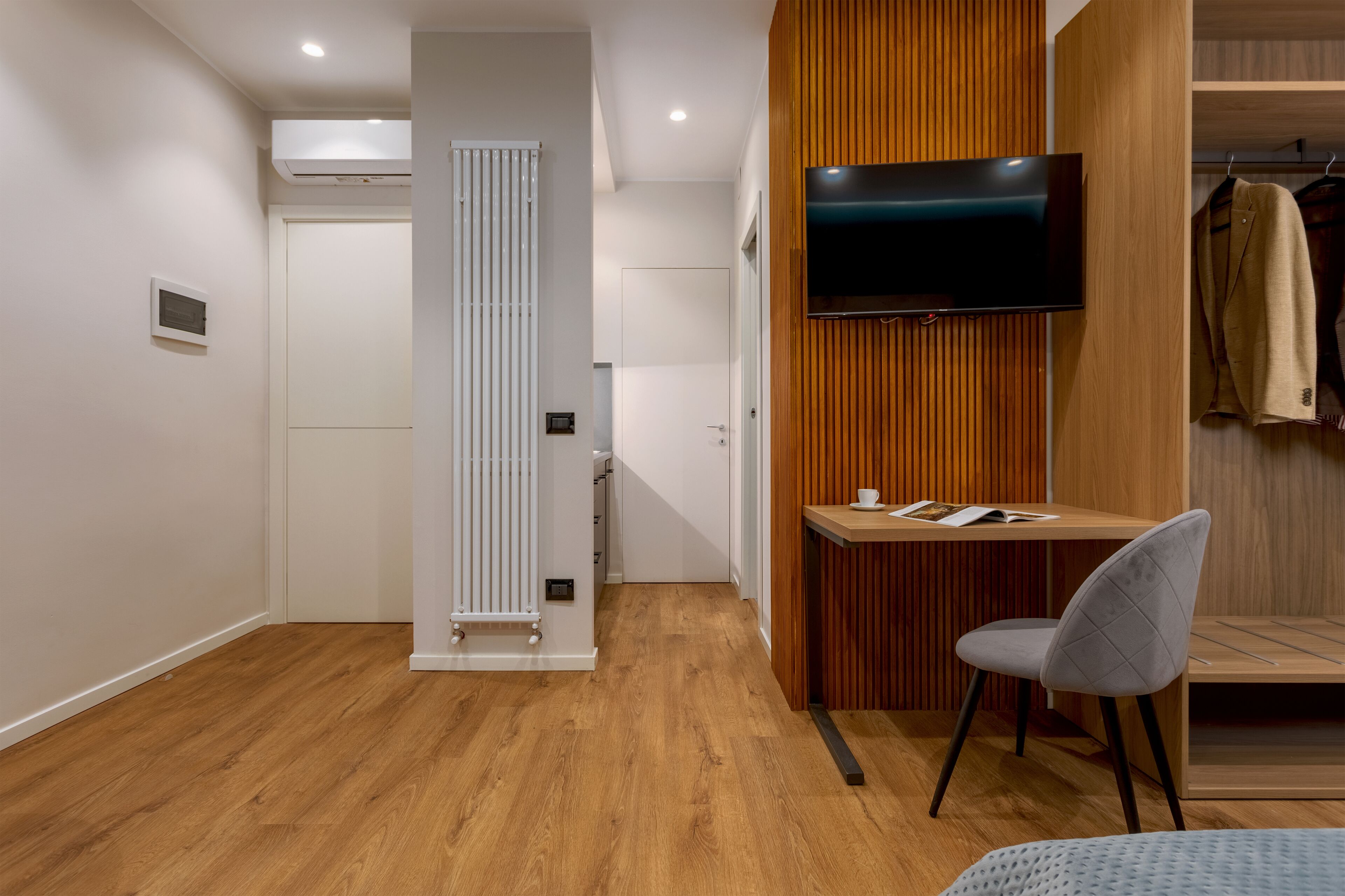 Une chambre moderne avec un plancher en bois, un radiateur blanc, un bureau avec une chaise, un téléviseur fixé au mur et un portant à vêtements.