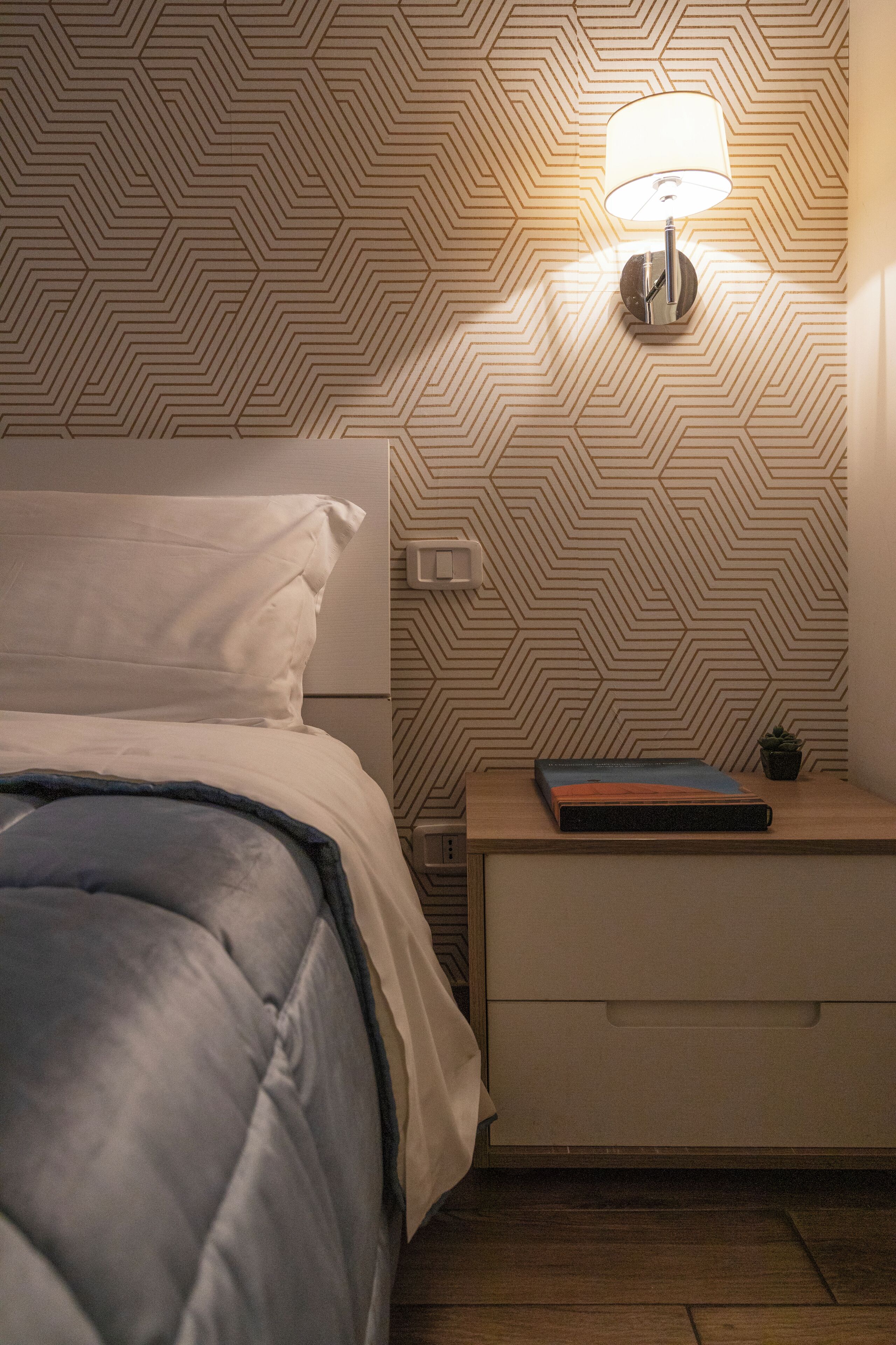 A neatly made bed with a white pillow and a blue blanket, a bedside table with a lamp, a book, and a small plant.