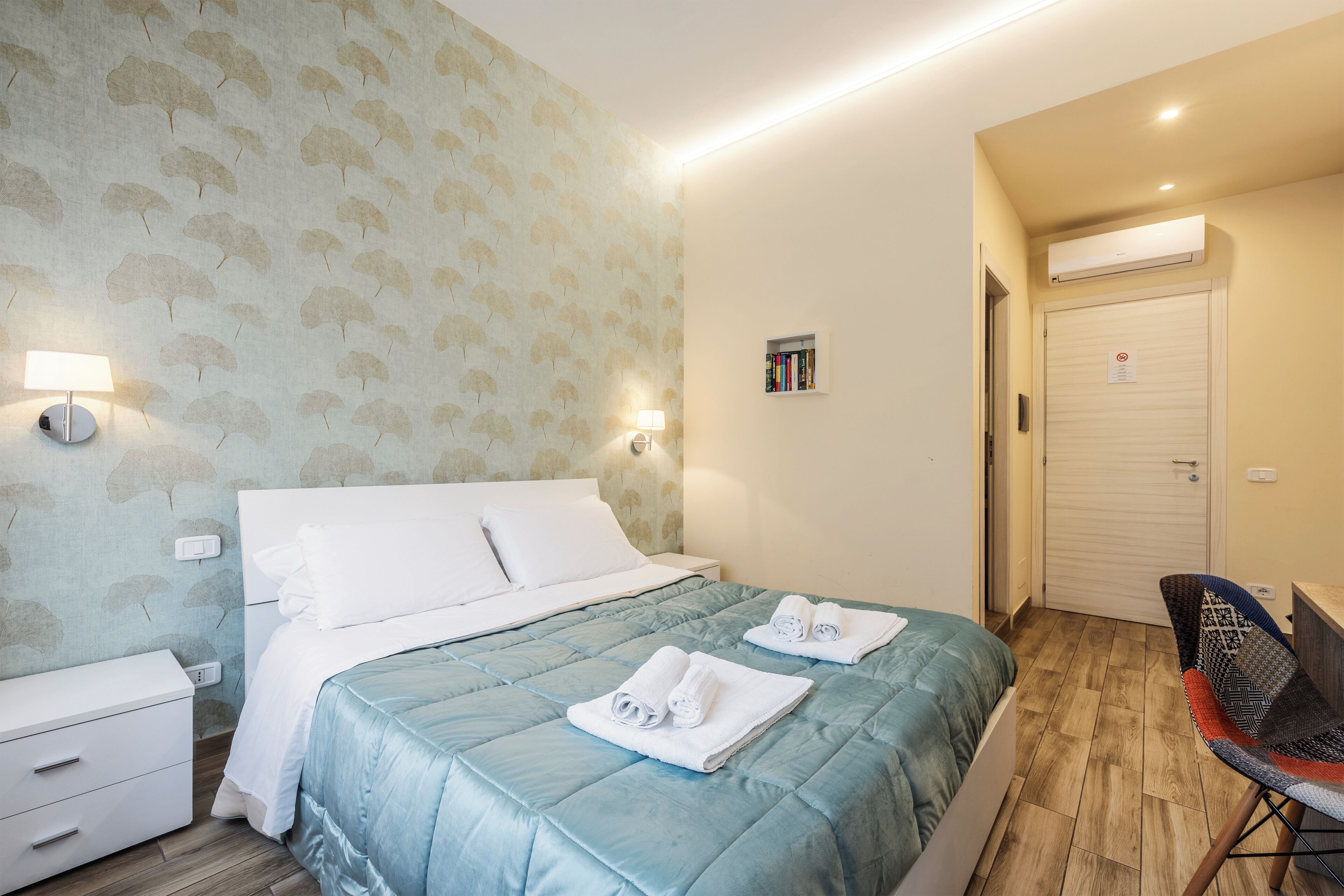 Habitació d'hotel amb un llit, una tauleta de nit, una làmpada i una petita estant amb llibres.