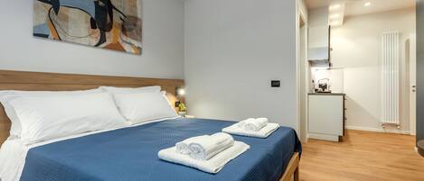 A hotel room with a large bed, two pillows, a blue bedspread, and two rolled towels on the bed.