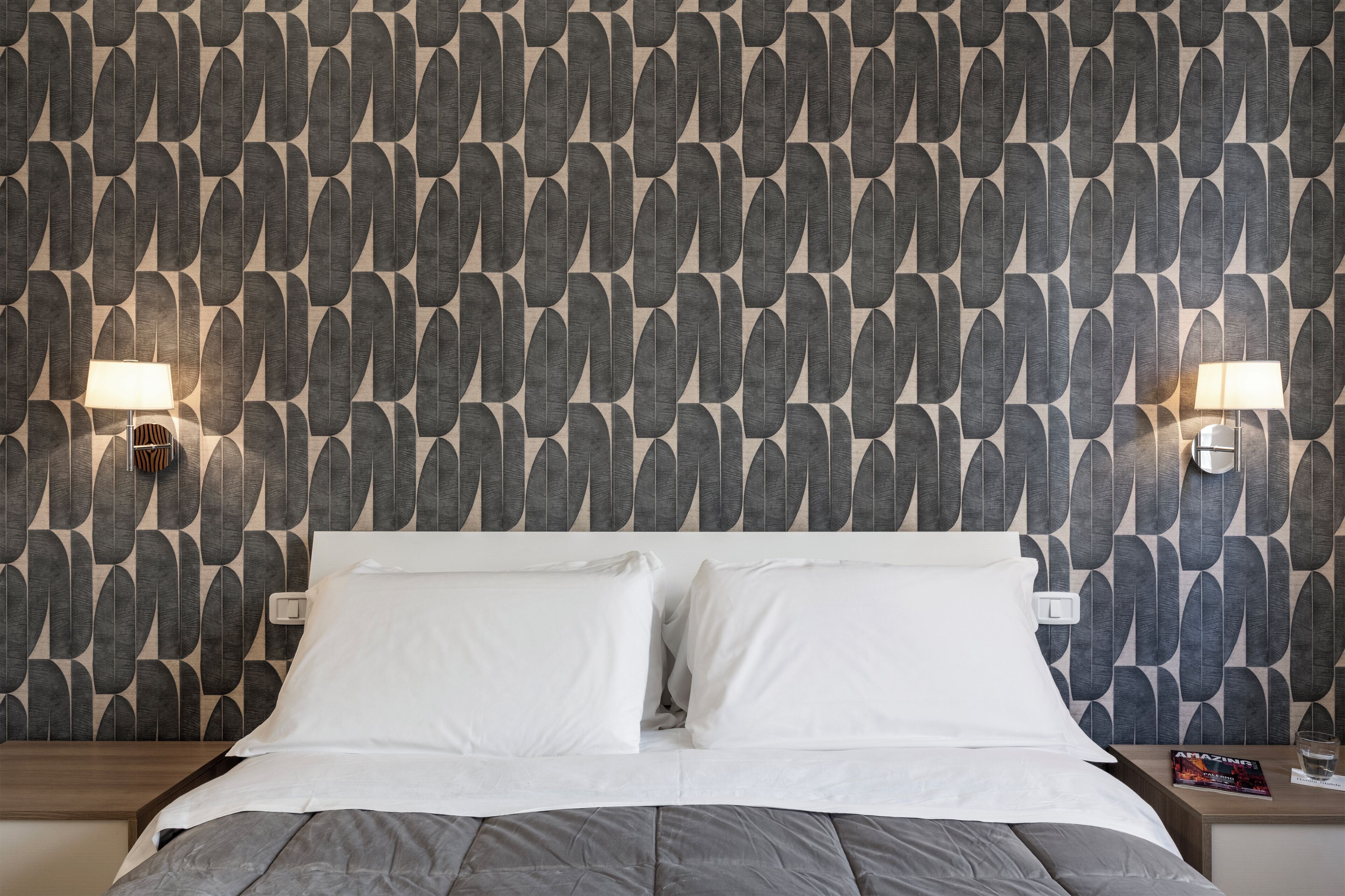 A hotel room with a patterned wallpaper, a bed with white linens, two bedside lamps, and a magazine on the nightstand.