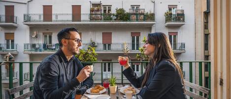 Un couple est assis à une table en plein air, devant un immeuble à plusieurs étages doté de balcons.