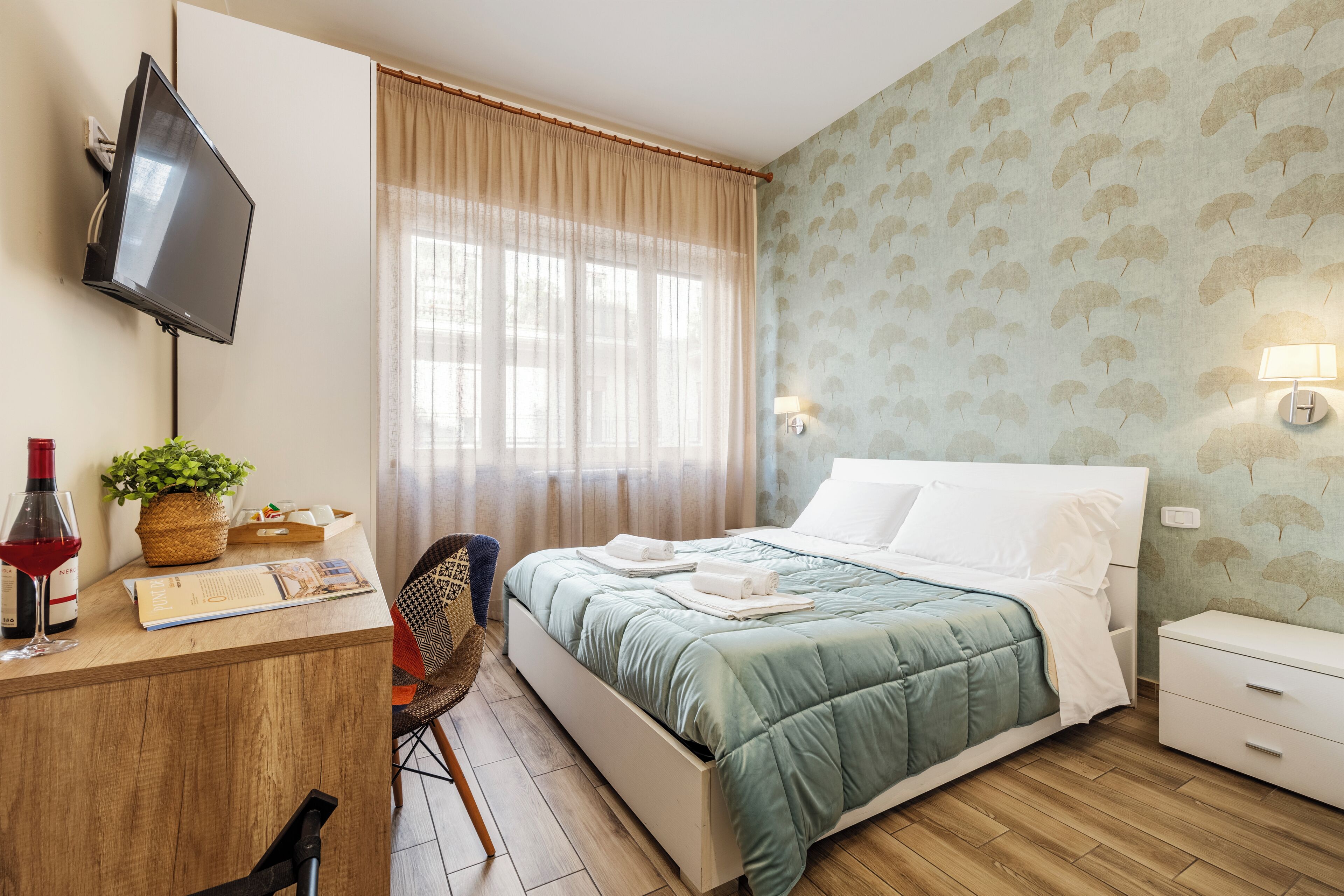 Habitació d'hotel amb llit, escriptori amb ordinador, televisió, finestra amb cortines i paret amb un dibuix de fulles.