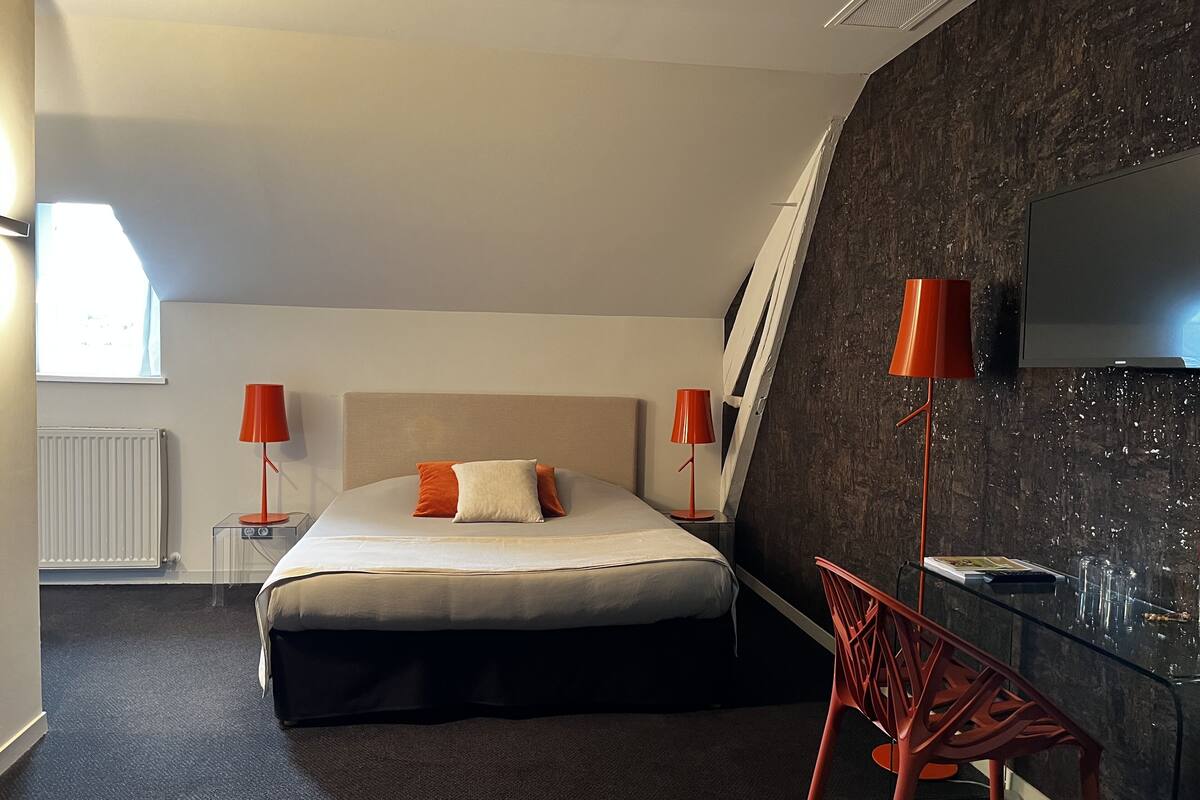 Habitación de hotel moderna con una cama grande, dos lámparas rojas, un escritorio de vidrio y un televisor montado en una pared oscura con textura.
