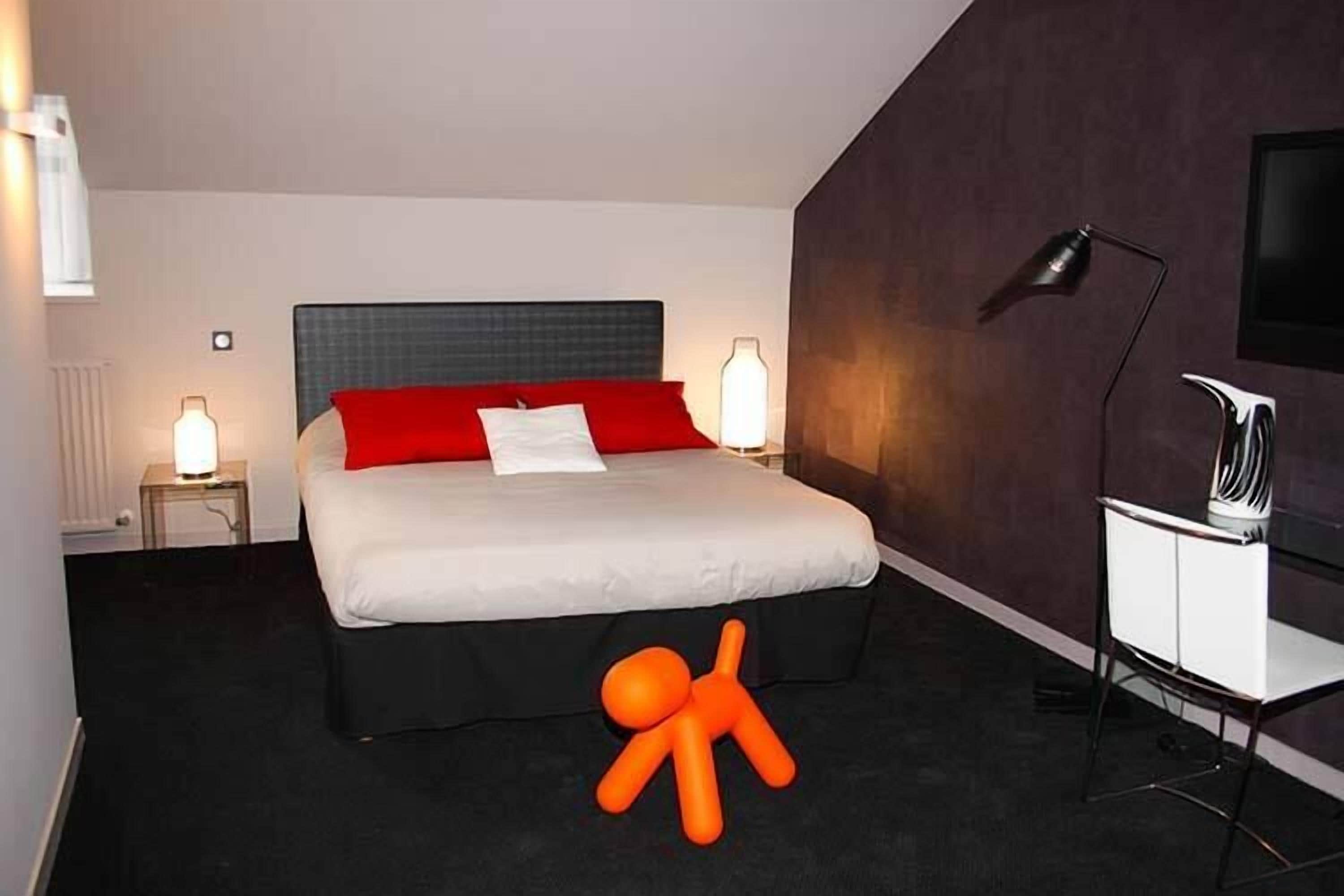 Ein modernes Hotelzimmer mit einem großen Bett, roter und weißer Bettwäsche, einem schwarzen Kopfteil, einem Schreibtisch mit Stuhl und einem Fernseher an der Wand.