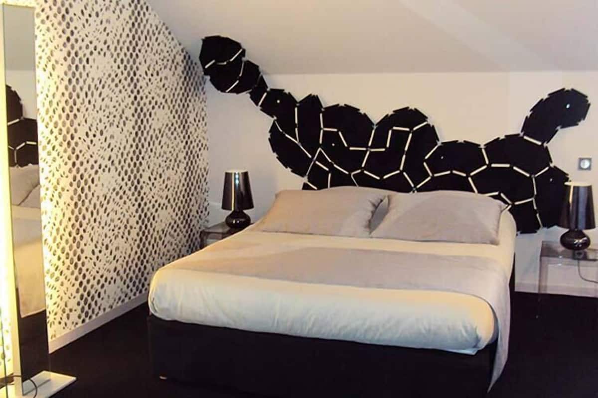 Un dormitorio moderno con un cuadro de arte geométrico, una cama con ropa blanca y una mesita de noche con una lámpara.