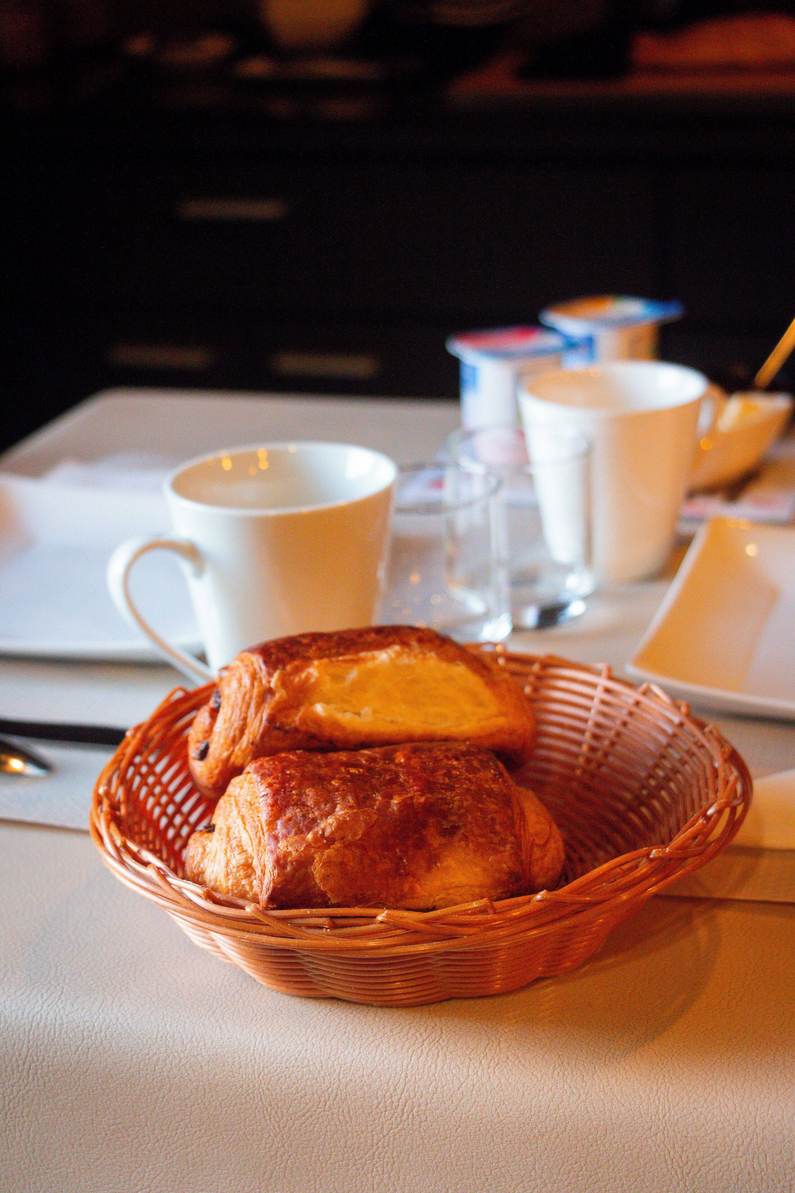 A wicker basket with pastries on a table, accompanied by white cups and a spoon.