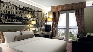 A hotel room with a large bed, a desk with a lamp, a television, and a balcony with a view of the city. - Mell Hotel (Trabzon)