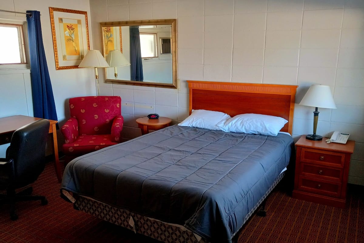 A hotel room with a bed, a desk, a chair, a bedside table, a lamp, and a mirror.