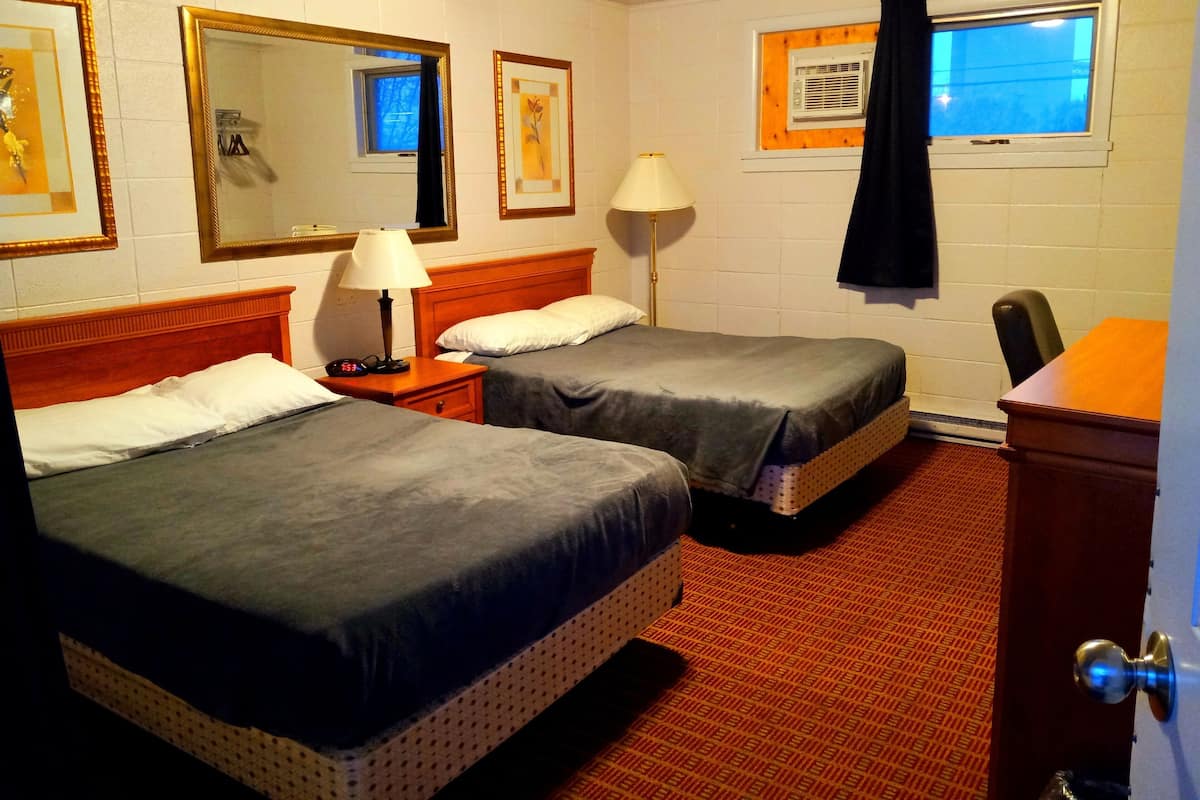 A hotel room with two beds, a nightstand with a lamp, a dresser, a window with a curtain, and a wall-mounted TV.