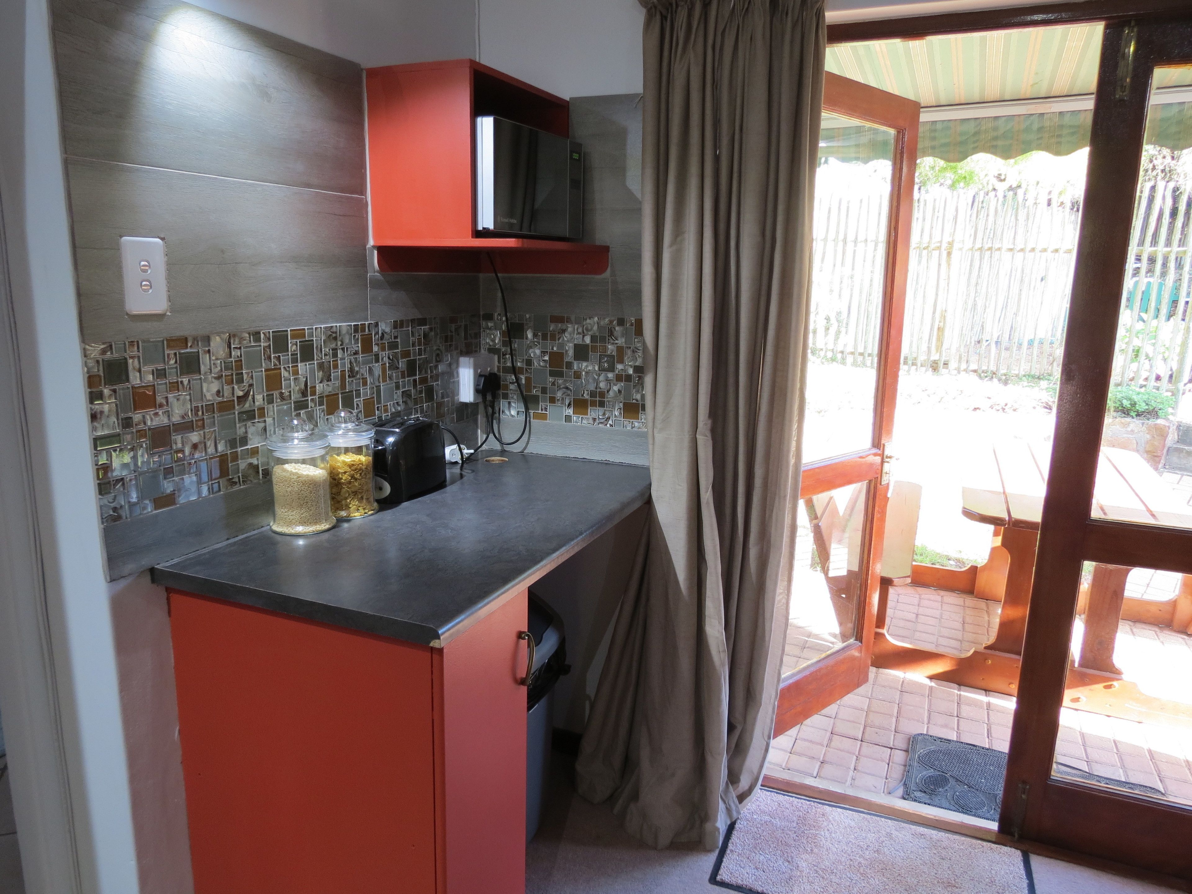 A compact kitchen with a microwave, toaster, and jars on the counter. A sliding door leads to an outdoor area.