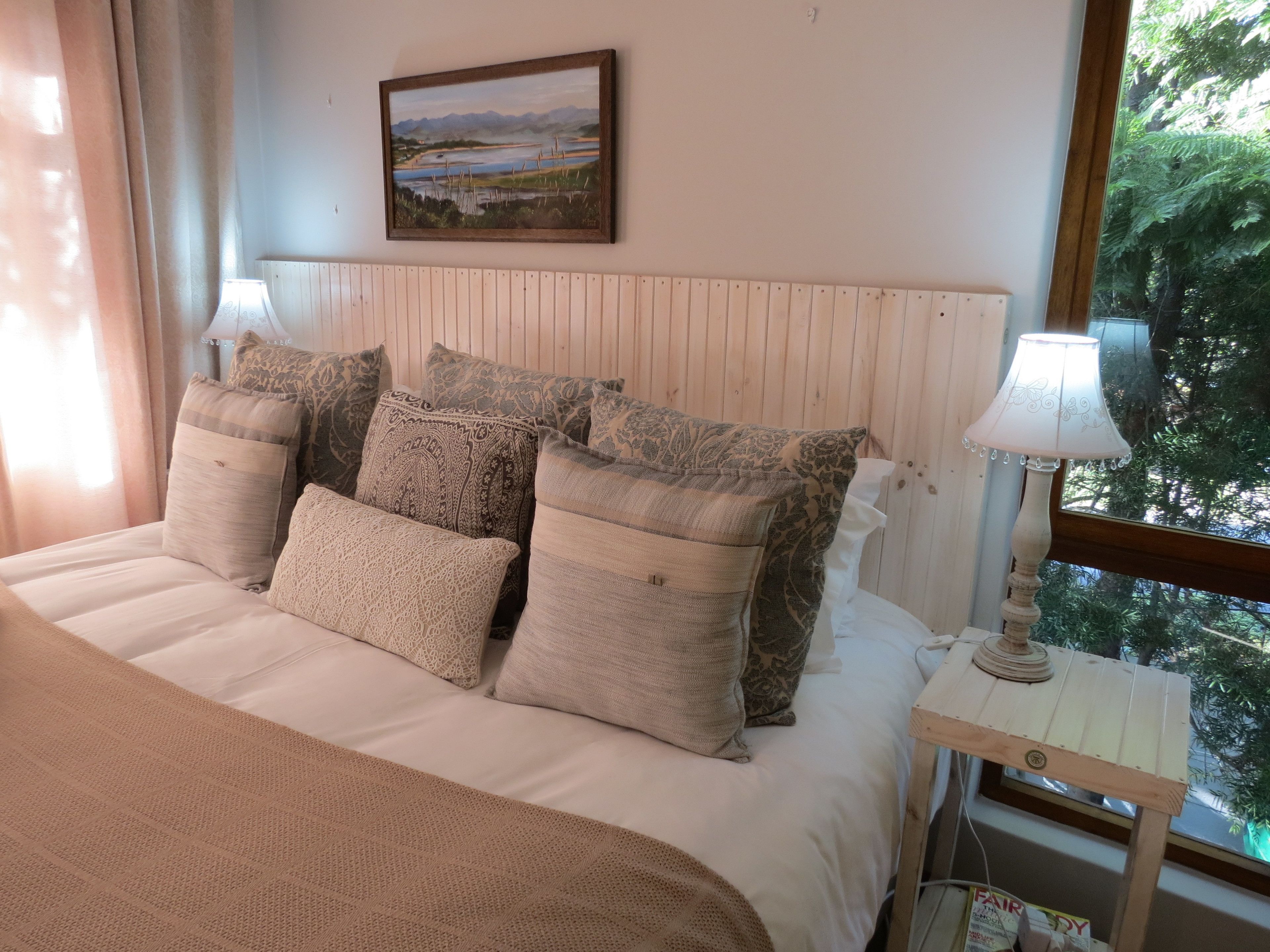 A bedroom with a bed, multiple pillows, a bedside table with a lamp, a painting on the wall, and a window with a view of trees.