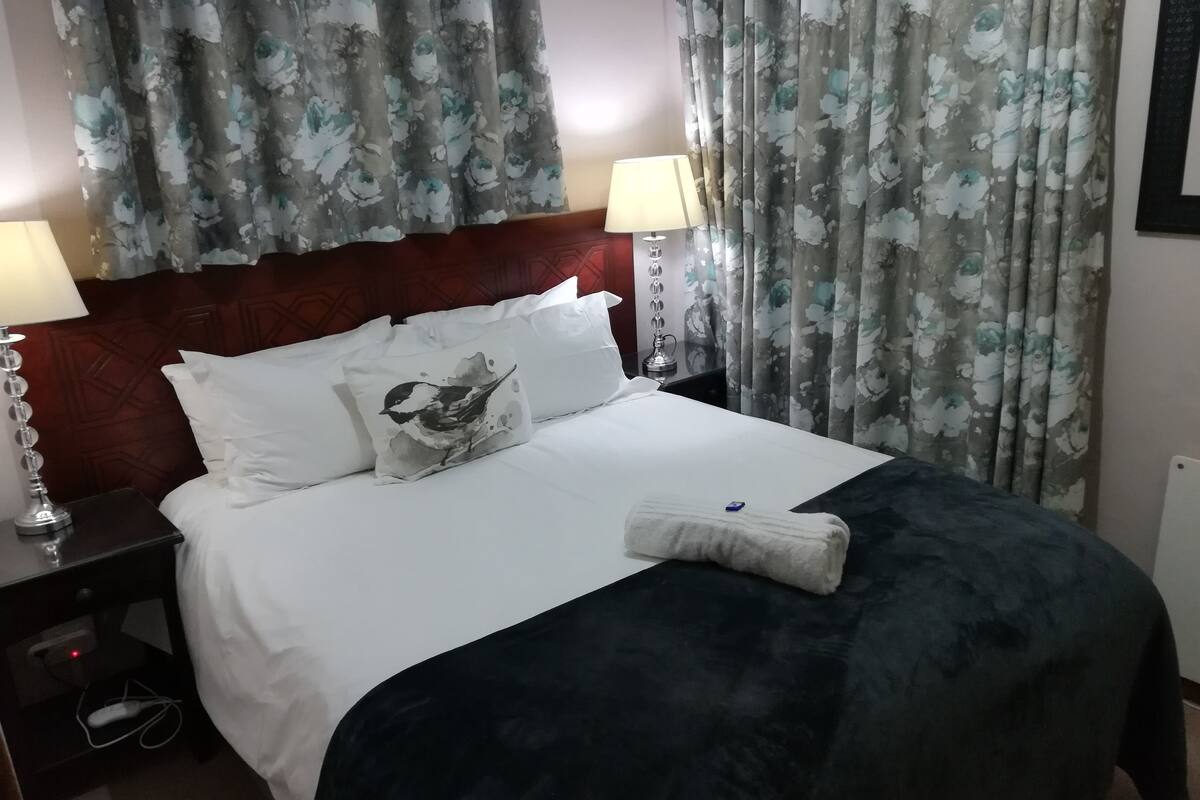 A neatly made bed with white linens and a dark blanket, flanked by two bedside tables with lamps, a red headboard, and floral curtains.
