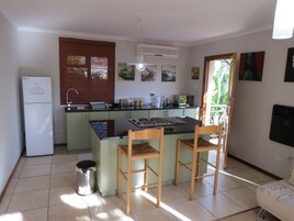 Cocina compacta con refrigerador, estufa y lavamanos. Hay dos sillas de madera y un basurero.