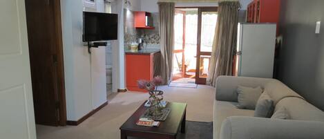 A compact living space with a sofa, a coffee table, a TV mounted on the wall, and a kitchenette area.