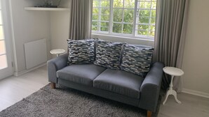 Ein Wohnzimmer mit grauem Sofa, gemusterten Kissen, einem Flachbildfernseher, einem Fenster mit Vorhängen und einem kleinen weißen Beistelltisch.