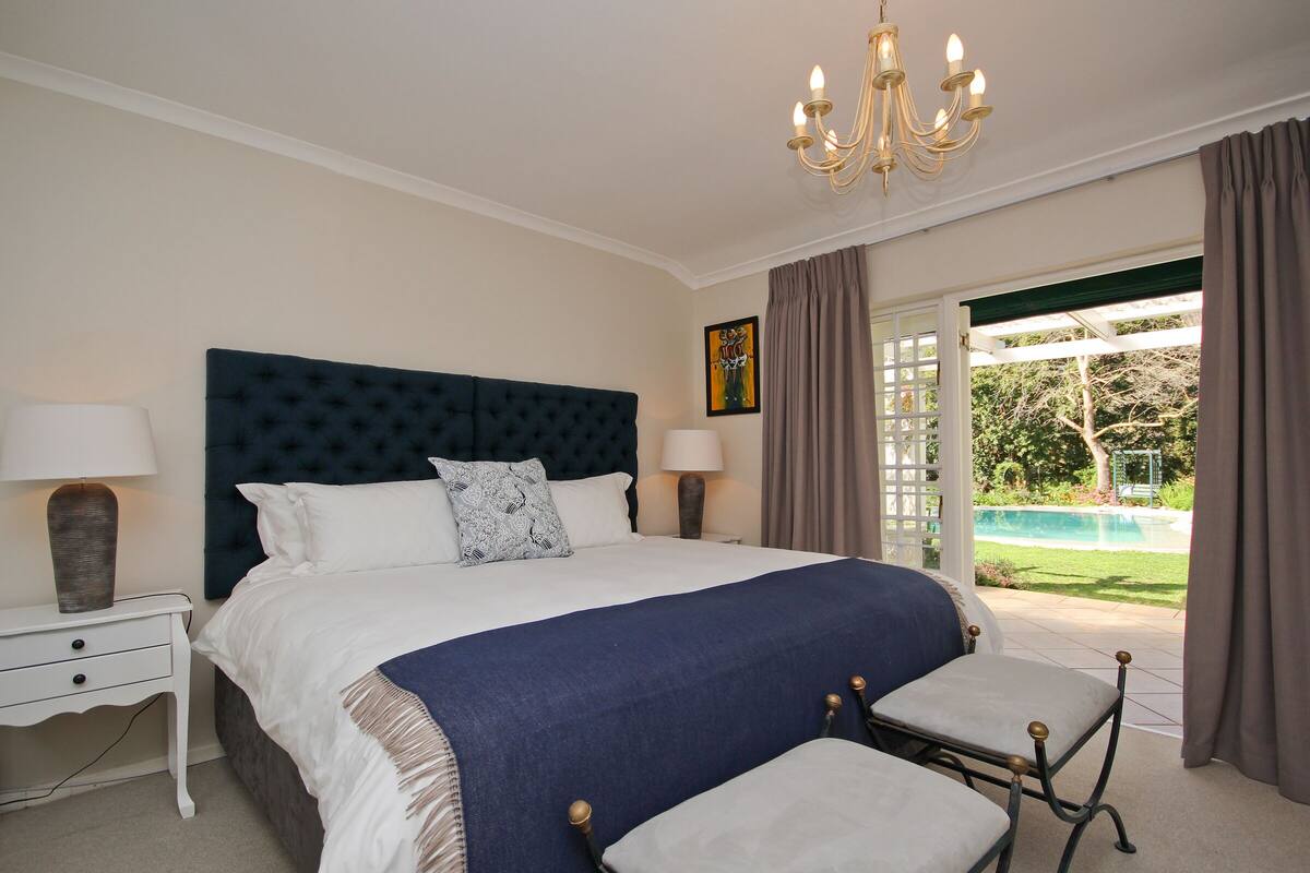 A bedroom with a large bed, bedside lamps, a chandelier, and a view of a swimming pool.