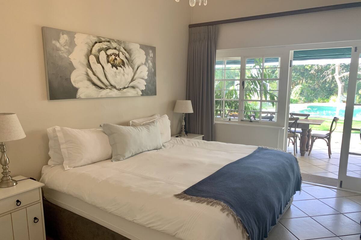 A bedroom with a large bed, a nightstand with a lamp, a painting on the wall, and a view of the outdoors through a sliding door.
