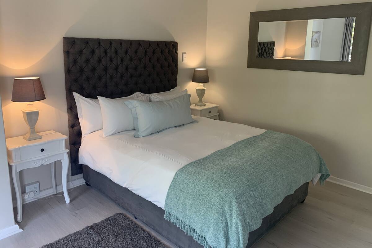 A neatly made bed with a tufted headboard, two bedside tables with lamps, a mirror on the wall, and a light-colored rug.