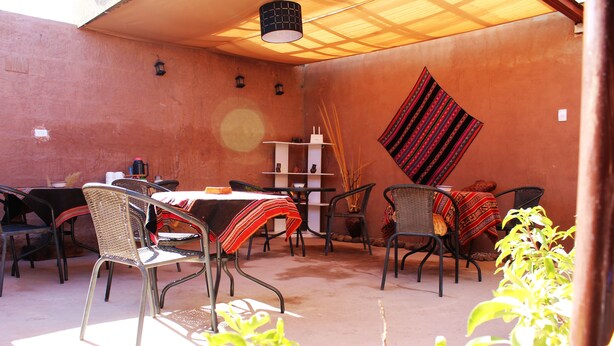An outdoor patio with metal chairs and tables, a woven chair, a hanging red and black textile, and a white shelf with decorative items. - Casa Sirius (San Pedro de Atacama)