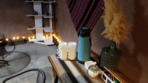 A cozy seating area with a wicker chair, a small table with a blue water bottle, white mugs, a glass jar, and a decorative vase with dried plants. - Casa Sirius (San Pedro de Atacama)